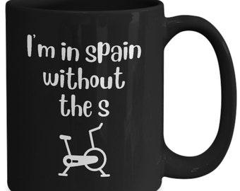 Peut inclure: Mug à café noir avec du texte blanc qui dit "Je suis en Espagne sans le s" et une silhouette blanche d'un vélo d'exercice stationnaire.