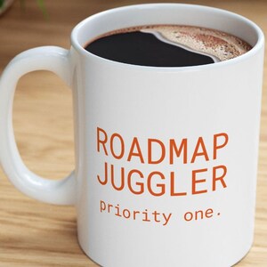 May include: A white ceramic mug with a large handle, filled with black coffee. The front of the mug features the orange text ROADMAP JUGGLER in a bold sans-serif font, followed by the smaller orange text priority one. in a simple font.