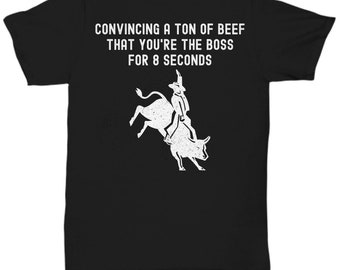 May include: Black t-shirt with white text that reads "Convincing a ton of beef that you're the boss for 8 seconds". A white silhouette of a cowboy riding a bucking bull is below the text.