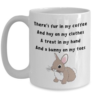 May include: White ceramic mug with a handle. The mug has the text "There's fur in my coffee, And hay on my clothes, A treat in my hand, And a bunny on my toes." A cartoon bunny illustration is also on the mug.