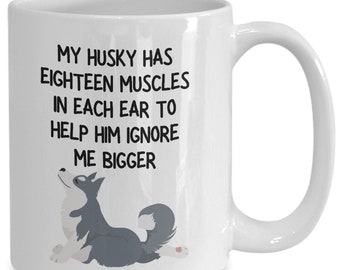 May include: White ceramic coffee mug with a black and white illustration of a husky dog. The text on the mug reads "My husky has eighteen muscles in each ear to help him ignore me bigger."