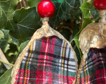 May include: Close-up of handmade Christmas ornaments. Each ornament features a plaid pattern in red, green, black, and white, with a gold-colored trim. The ornaments are topped with red beads and green glass accents, hanging among green leaves.