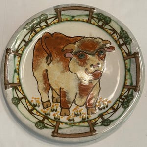 May include: Round ceramic plate featuring a brown and white bull illustration. The bull stands in a field of yellow grass, surrounded by a decorative border with green accents and brown framing. The plate has a light gray rim.