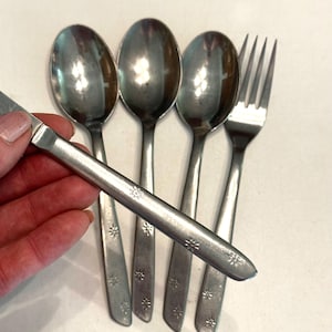 Puede incluir: Un juego de cubiertos antiguos, que incluye tres cucharas, un tenedor y un cuchillo. Los utensilios de acero inoxidable tienen un diseño de estrella en los mangos. Los cubiertos están dispuestos sobre una superficie blanca.