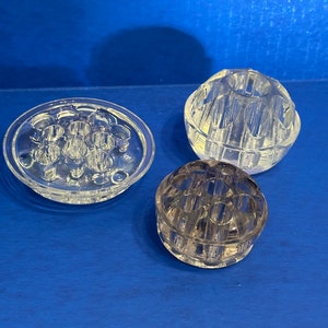 May include: Three clear glass flower frog bases. One is a round dish with multiple holes, another is a sphere with holes, and the last is a round, flat base with holes.