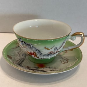 May include: A green and white teacup and saucer set. The cup and saucer feature a detailed dragon design in shades of gray, red, and gold. The cup has a gold handle and trim. The set is likely vintage or antique.