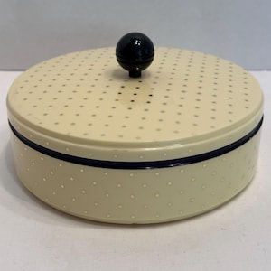 May include: A round, cream-colored lidded container featuring a repeating pattern of small, light-colored dots. The lid is topped with a smooth, black spherical knob, and a thin, dark horizontal band encircles the side of the container base.