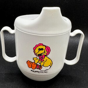 Puede incluir: Vaso para sorber de plástico blanco con dos asas y tapa. El vaso presenta una colorida ilustración de Big Bird de Barrio Sésamo, con un gorro y un lazo rosa. El texto "MUPPETS INC." está impreso debajo.