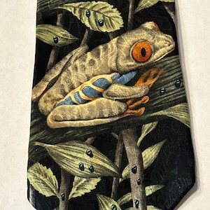 Puede incluir: Una corbata de seda negra con un patrón repetido de hojas verdes, ramas marrones y una rana verde y naranja con ojos rojos.