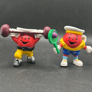 Può includere: Due piccole figure rosse dei cartoni animati. Una figura solleva un bilanciere con pesi marroni, indossa pantaloni gialli e scarpe nere. L'altra figura tiene in mano una racchetta da tennis verde con una palla viola, indossa una camicia gialla, pantaloncini blu e scarpe bianche.