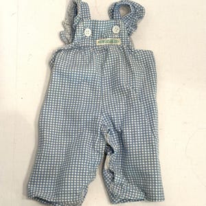 May include: A light blue and white checkered overall featuring ruffled shoulder straps, two white buttons on the front bib, and a small rectangular label with text centered below the buttons. The garment has a straight-leg design.