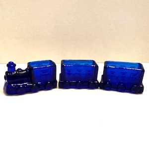 May include: A cobalt blue glass train set, featuring a locomotive and two train cars. The train has detailed features, including wheels and a smokestack. The set is displayed on a white surface, showcasing the vibrant color of the glass.