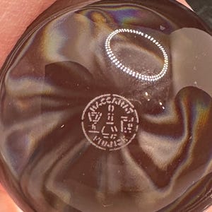 May include: A round, dark brown glass object with a smooth, iridescent surface featuring swirling patterns. A circular stamp is etched into the center, containing the text "VECCHIA" at the top and "MURANO" at the bottom, with a stylized tower icon in the middle.
