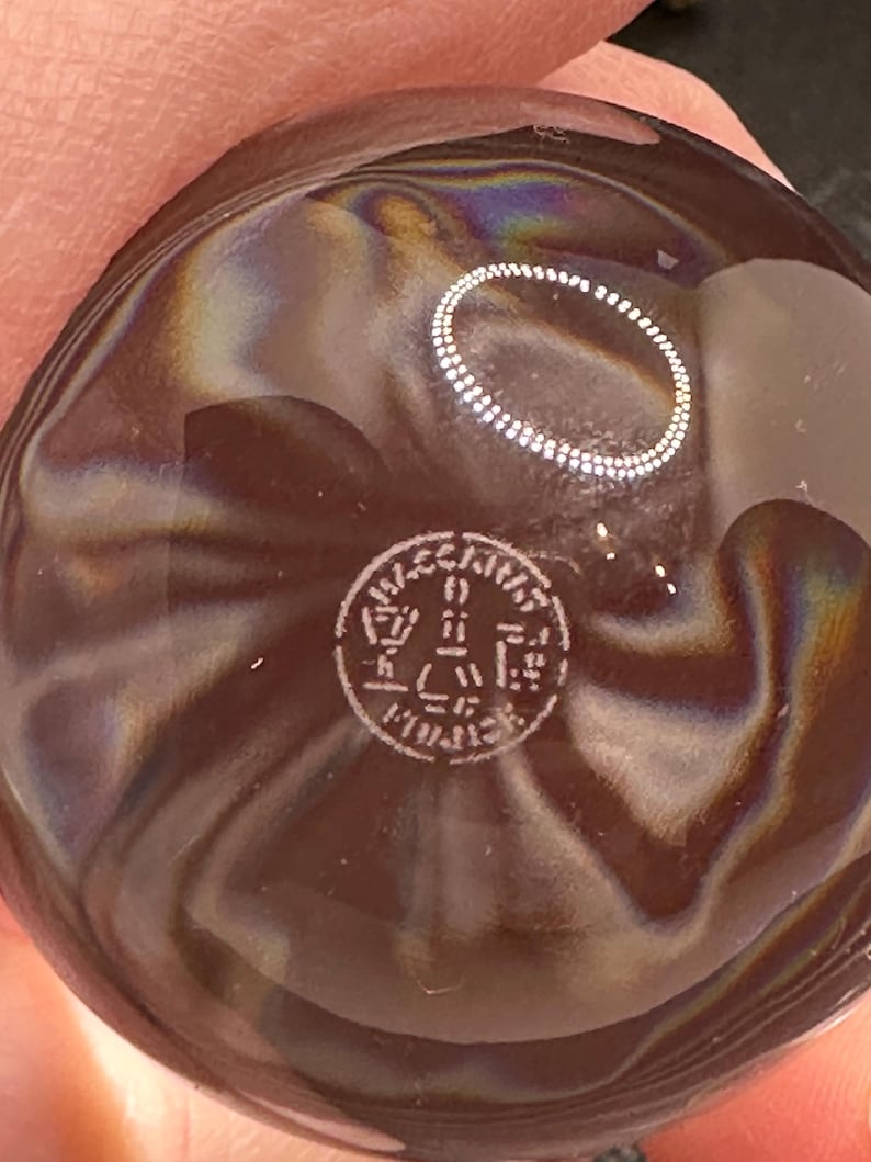 May include: A round, dark brown glass object with a smooth, iridescent surface featuring swirling patterns. A circular stamp is etched into the center, containing the text "VECCHIA" at the top and "MURANO" at the bottom, with a stylized tower icon in the middle.