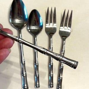 Puede incluir: Un juego de cubiertos de acero inoxidable con un diseño de mango estilo bambú. El juego incluye dos cucharas y dos tenedores. Los cubiertos se muestran sobre una superficie clara.