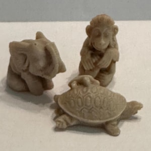 Op de afbeelding: Drie kleine, bruinbeige dierenbeeldjes: een olifant, een aap en een schildpad. De olifant heeft grote oren en een slurf. De aap zit met zijn handen bij zijn gezicht. De schildpad heeft een getextureerd schild.