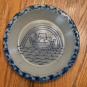 Puede incluir: Un plato de cerámica azul y blanco con una ilustración azul y blanco de un barco con un arcoíris encima. El barco está lleno de objetos redondos y tiene una persona de pie en la parte delantera del barco.