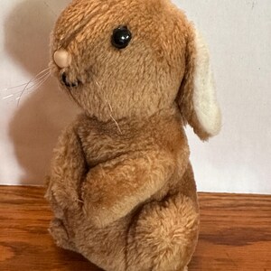 Puede incluir: Un juguete de peluche de conejo vintage en tonos marrones. El conejo tiene una cabeza redonda, orejas largas y una pequeña nariz rosada. Tiene ojos negros y bigotes largos y finos. El conejo está sentado.