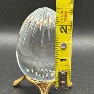 May include: A clear, egg-shaped glass paperweight with vertical cut facets, resting on a gold-toned metal tripod stand with ornate feet. A yellow measuring tape placed next to the object indicates a height of approximately 2.5 inches.