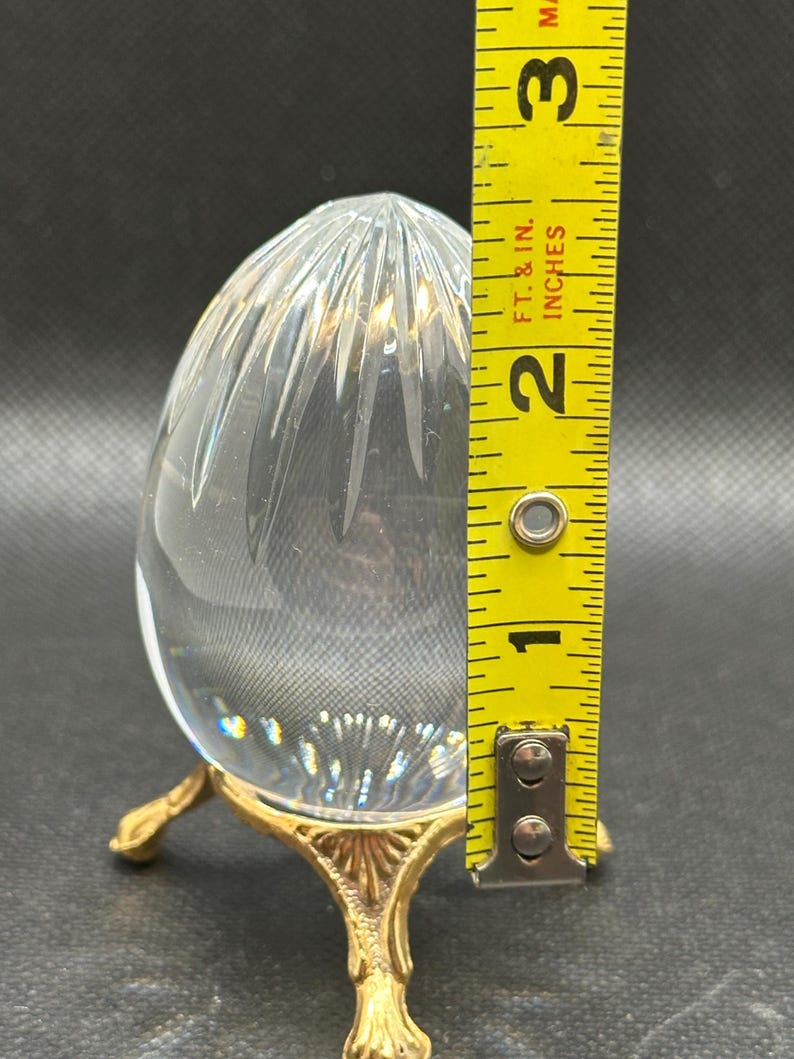 May include: A clear, egg-shaped glass paperweight with vertical cut facets, resting on a gold-toned metal tripod stand with ornate feet. A yellow measuring tape placed next to the object indicates a height of approximately 2.5 inches.