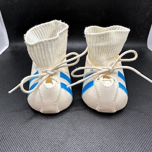 May include: A pair of baby shoes with white uppers and blue stripes. The shoes have white laces and a ribbed, sock-like cuff. The shoes are designed to look like miniature sneakers.