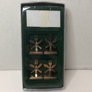 May include: Set of four gold-tone metal place card holders in a green box. Each holder features a fork, knife, and spoon design.