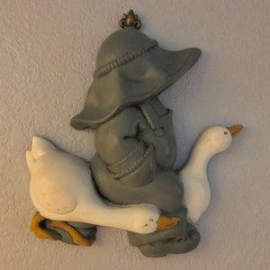 May include: A gray figurine of a person wearing a hat and riding a white goose. The figurine is made of plastic and has a rustic, vintage look.