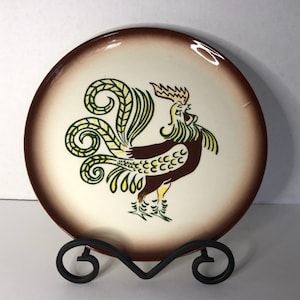 Peut inclure: Une assiette en céramique avec un motif de coq. L'assiette a un centre crème avec un bord marron. Le coq est marron, jaune et vert. L'assiette est présentée sur un présentoir en métal noir.