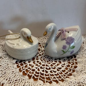 May include: Two white porcelain swan figurines. One swan has a pink ribbon and purple flowers. The other swan has a gold beak and is in a nest-like base.