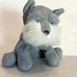 May include: A plush gray Schnauzer dog toy with a white fluffy beard and eyebrows. The toy has small, floppy ears and a black nose. The dog is sitting upright, with its front paws visible. The toy is made of soft, textured material.