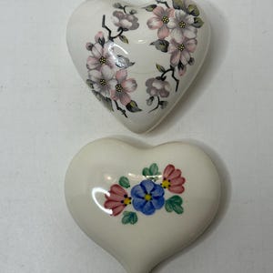 以下が含まれることがあります： 花柄の白いセラミック製のハート型のオーナメント2つ。上のオーナメントはピンクとグレーの花、下のオーナメントはピンク、ブルー、グリーンの花が描かれている。