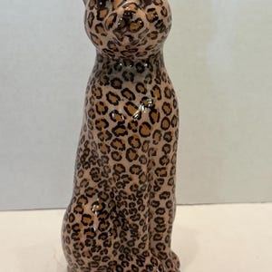 May include: A ceramic figurine of a seated leopard with a glossy finish. The sculpture features a light tan base color covered in a pattern of dark brown and black leopard spots across the entire body, including the head, ears, and torso.