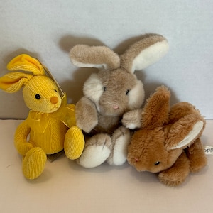 May include: Three plush bunny rabbits, one yellow, one brown, and one light brown. The rabbits are sitting on a white surface.