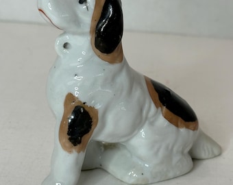 May include: A white porcelain figurine of a dog sitting with its head tilted up. The dog has black and brown markings on its body.