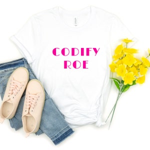 Peut inclure: T-shirt blanc avec le texte "CODIFY ROE" en lettres roses. Le t-shirt est associé à un jean bleu et des baskets roses.