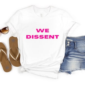 Peut inclure: T-shirt blanc avec le texte "WE DISSENT" imprimé en rose vif.