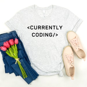 Op de afbeelding: Een grijs t-shirt met de tekst "<CURRENTLY CODING/>" in zwart gedrukt.
