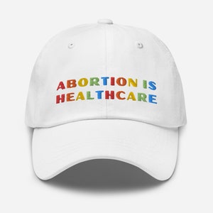 Peut inclure: Casquette de baseball blanche avec le texte "ABORTION IS HEALTHCARE" brodé en arc-en-ciel.