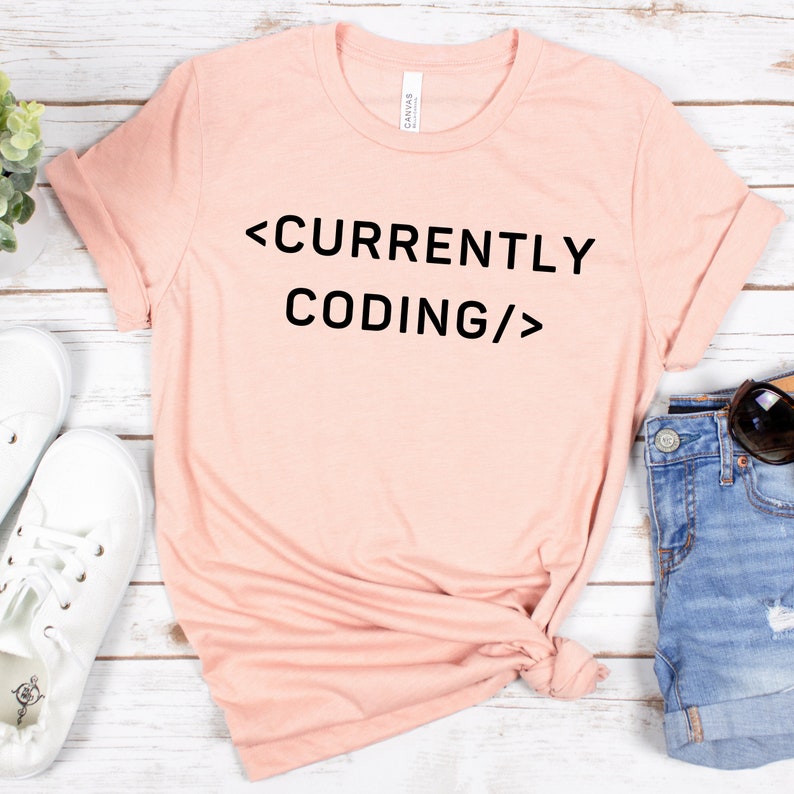 Op de afbeelding: Een lichtroze T-shirt met de tekst "<CURRENTLY CODING/>" in zwart gedrukt.