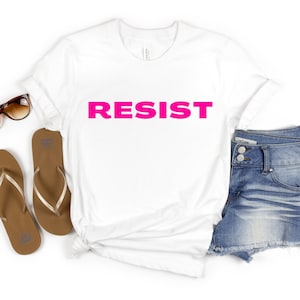 Peut inclure: T-shirt blanc avec le mot "RESIST" imprimé en rose vif.