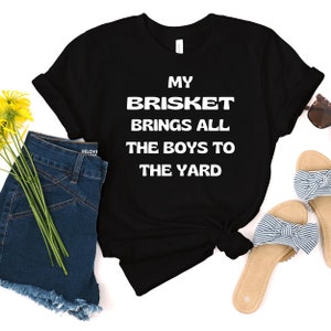 May include: Black t-shirt with white text that reads "My Brisket Brings All The Boys To The Yard". The shirt is laid out on a white surface with a pair of blue denim shorts, yellow flowers, and a pair of sandals.