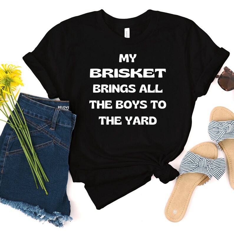 May include: Black t-shirt with white text that reads "My Brisket Brings All The Boys To The Yard". The shirt is laid out on a white surface with a pair of blue denim shorts, yellow flowers, and a pair of sandals.