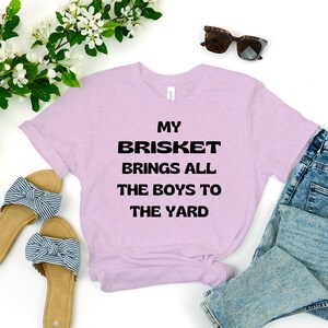 May include: A light purple t-shirt with the text "My Brisket Brings All The Boys To The Yard" printed in black.