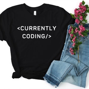 Op de afbeelding: Zwart T-shirt met witte tekst die "<CURRENTLY CODING/>" luidt. Het shirt ligt op een wit oppervlak met een blauwe spijkerbroek en roze bloemen.