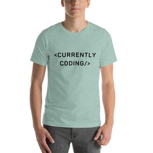 Op de afbeelding: Een lichtblauw T-shirt met de tekst "<CURRENTLY CODING/>" in zwart.