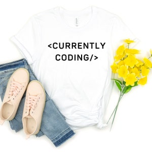 Op de afbeelding: Wit T-shirt met de tekst "<CURRENTLY CODING/>" in het zwart gedrukt.