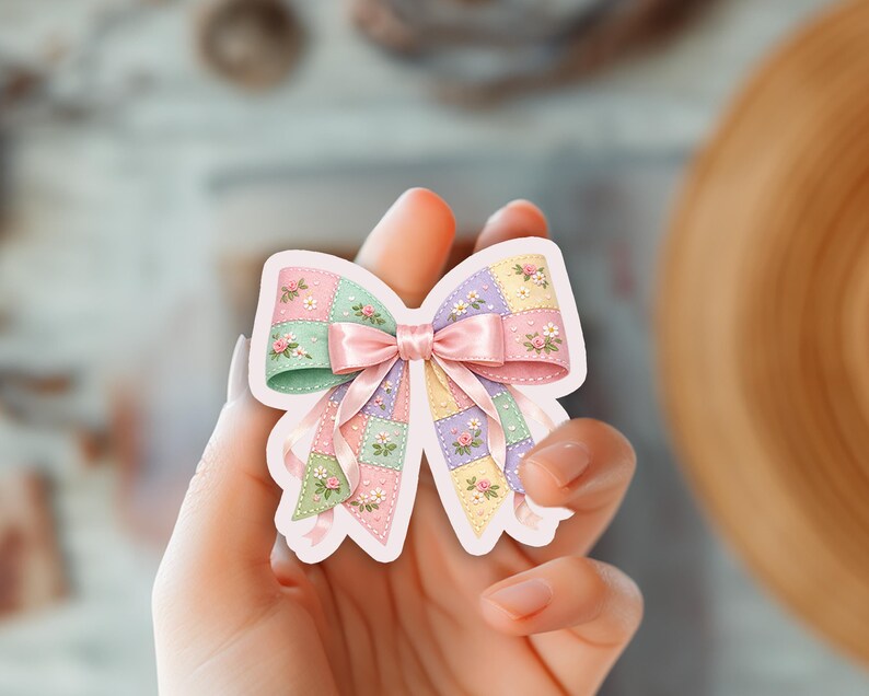 May include: A pastel-colored sticker in the shape of a bow. The bow features a patchwork design with pink, green, and purple squares, adorned with small floral accents. A pink satin ribbon is tied in the center, adding a delicate touch.