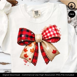 Puede incluir: Camiseta blanca de manga larga con un gran lazo decorativo. El lazo tiene patrones de cuadros rojos y negros y cuadros rojos y blancos. El lazo también tiene cinta beige con hojas de arce naranjas. La camiseta tiene una etiqueta Comfort Colors.