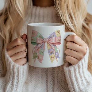 May include: White ceramic mug featuring a pastel patchwork bow design with pink, green, yellow, and purple squares. The bow has a pink ribbon in the center and floral accents. A charming and decorative coffee mug.