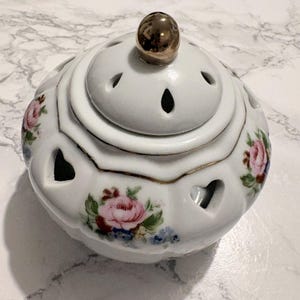 Peut inclure: Brûleur d'encens en céramique blanche avec un couvercle et un bouton doré. Le brûleur présente des découpes en forme de cœur et est décoré de motifs floraux roses et bleus et de bordures dorées. Un objet décoratif.